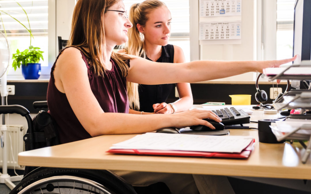 Zwei Frauen, wovon eine im Rollstuhl ist, sitzen an einem Schreibtisch und schauen auf eine Computermonitor