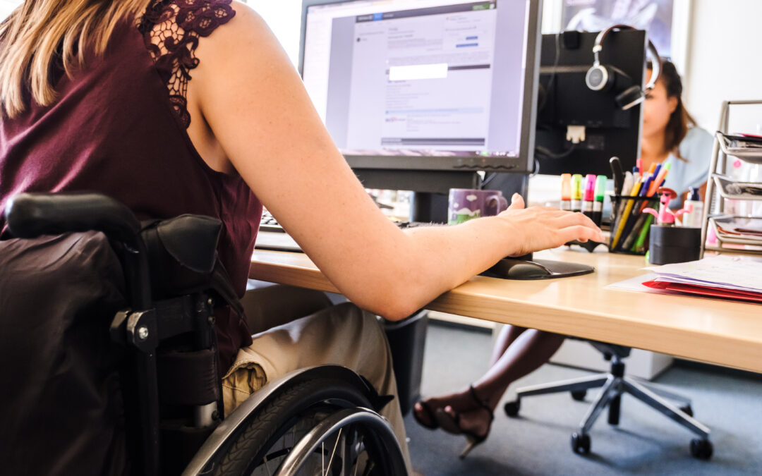 Zwei Frauen sitzen in einem Büro an Computern gegenüber. Eine von ihnen sitzt im Rollstuhl.