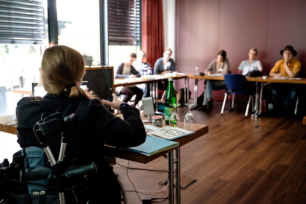 Eine Workshop-Geberin im Rollstuhl gibt einen Workshop. Sie sitzt vor einer Gruppe Menschen, die zuhören und mitschreiben an Tischen vor ihr.