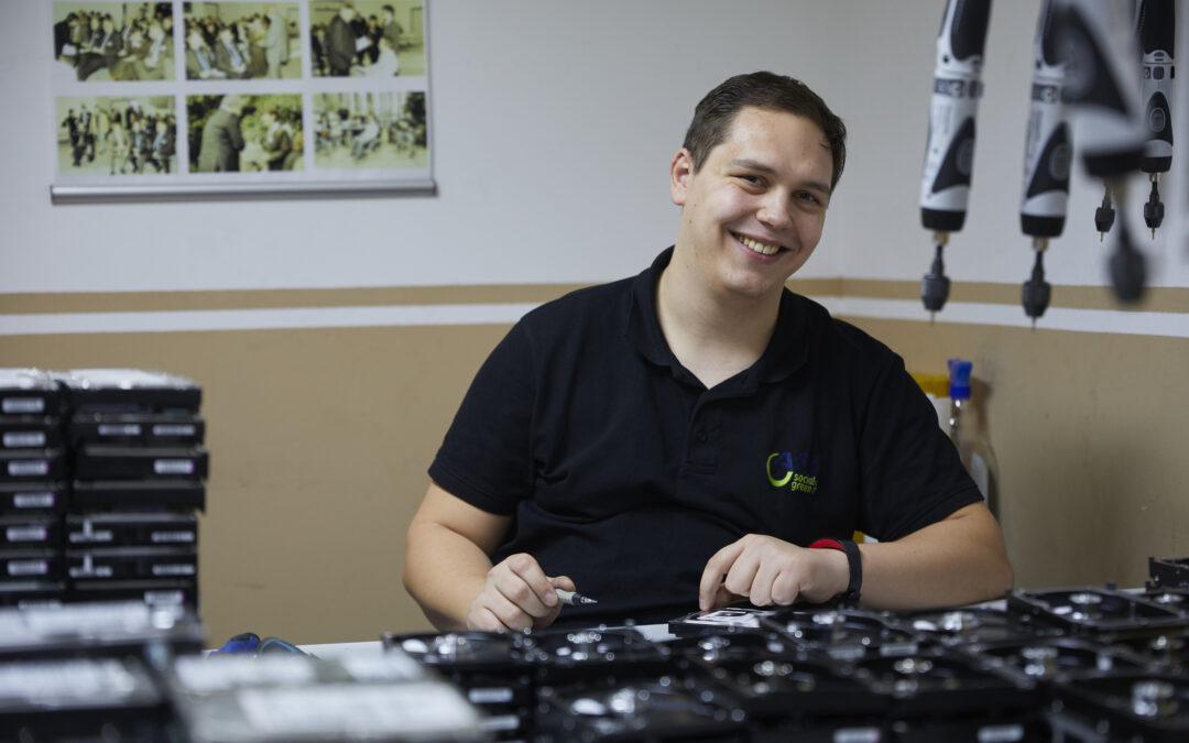 Ein junger Mann sitzt an einem Tisch und lächelt in die Kamera. Er schraubt an schwarzen technischen Geräten herum