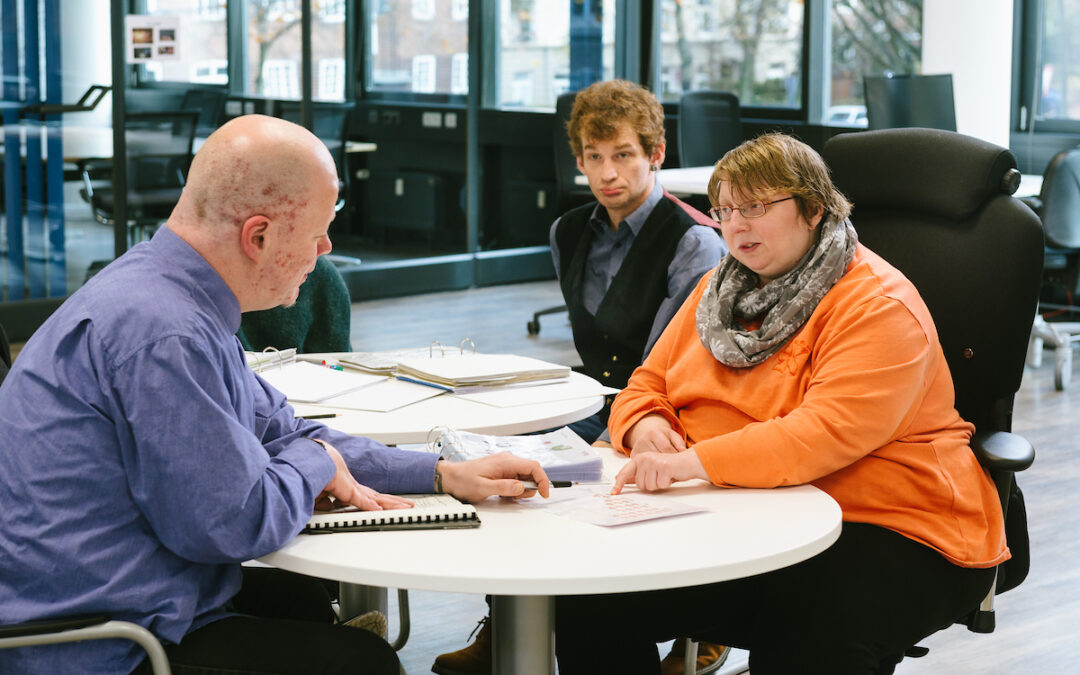 Drei Auszubildende sitzen an einem Tisch, auf dem Ordner und Zettel liegen, und üben eine Gesprächssituation.