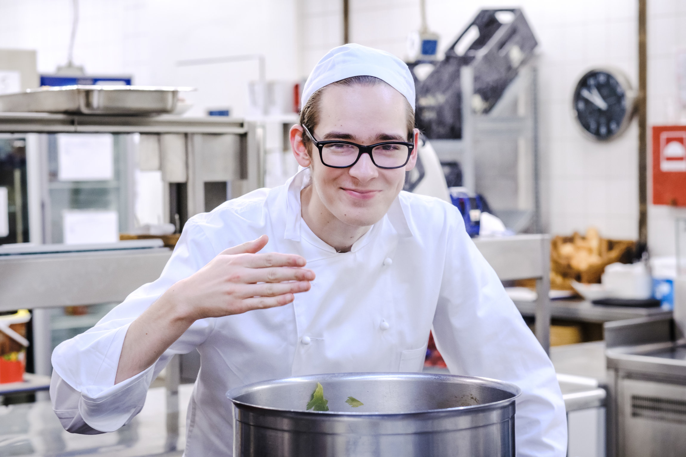 Ein weißer Mann mit Lernschwierigkeiten steht in einer Küche und schmeckt Essen ab. Er trägt eine weiße Kochschürze und eine weiße Kochmütze