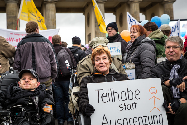 Bild von behinderten Demonstrierenden vor dem Brandenburger Tor. Sie halten ein Plakat in der Hand auf dem steht: Teilhabe statt Ausgrenzung.