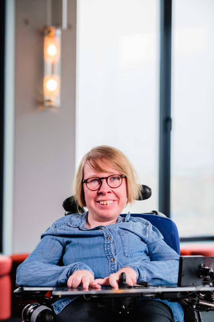 Eine weiße Frau mit blonden kinnlangen Haaren und Brille lächelt in die Kamera.