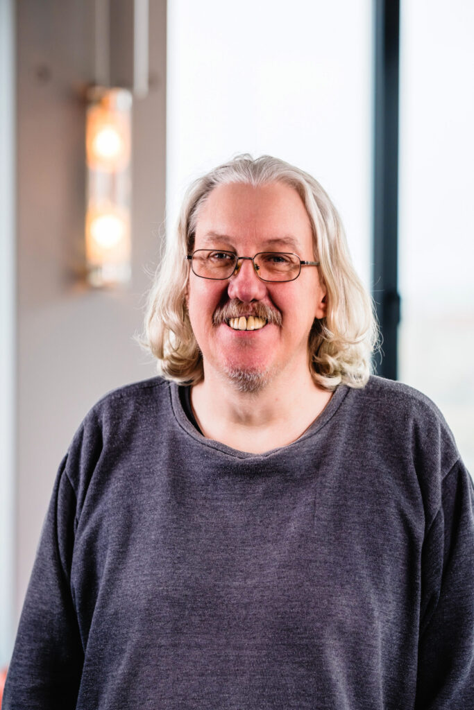 Ein weißer Mann mit grauem schulterlangem Haar, Schnurrbart und Brille lächelt in die Kamera.
