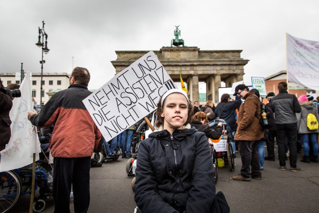 Ein Mensch mit Rollstuhl ist auf einer Demo vor dem Brandenburger Tor. Er hat ein Schild auf dem steht: Nehmt uns unsere Assistenz nicht weg