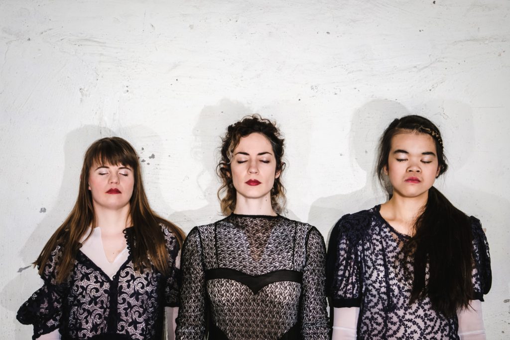 Drei weiblich gelesene Personen stehen vor einer Wand.
