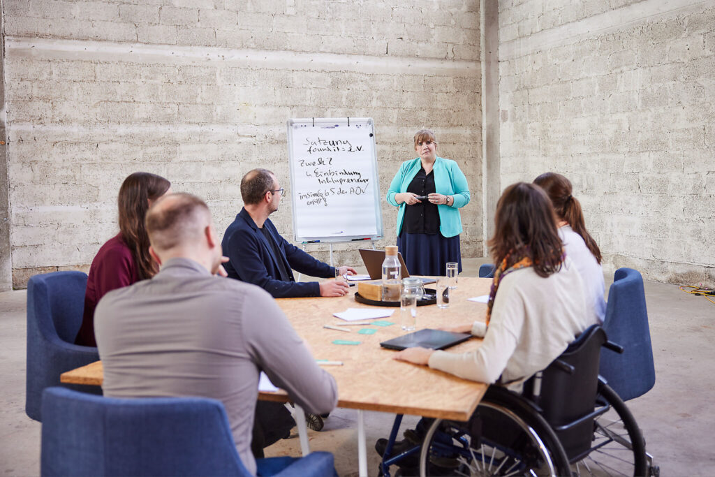 Aktuelles Bild: Fünf Menschen mit und ohne behinderung sitzen um einen Tisch. Vor ihnen steht eine Frau und hält einen Workshop.