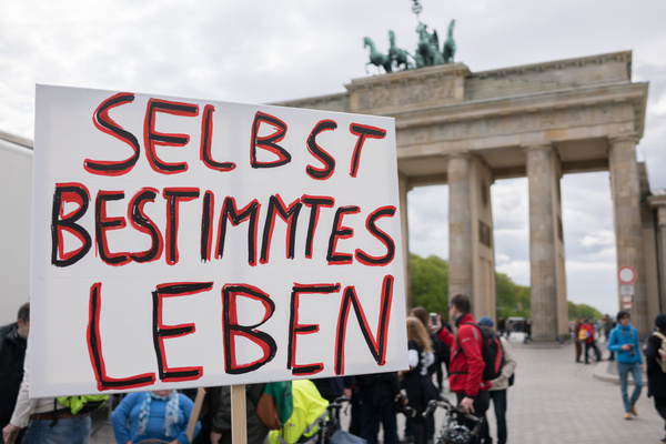 Ein Schild auf dem steht: Selbstbestimmtes Leben. Im Hintergrund ist das Brandenburger Tor