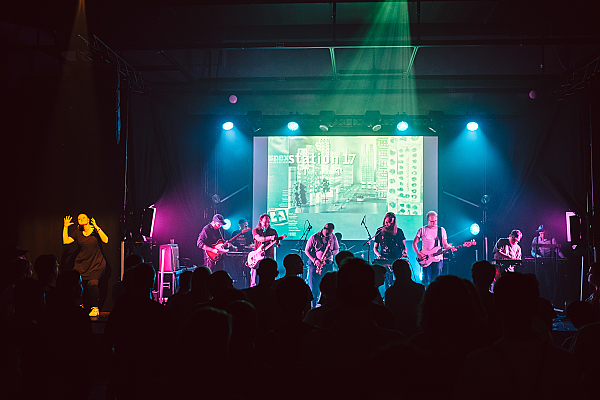 Eine Bühne und davor eine Menschenmenge. Auf der Bühne steht eine Gruppe von Musiker*innen. Sie spielen Bass, E-Gitarre, Saxophon und Keybord.